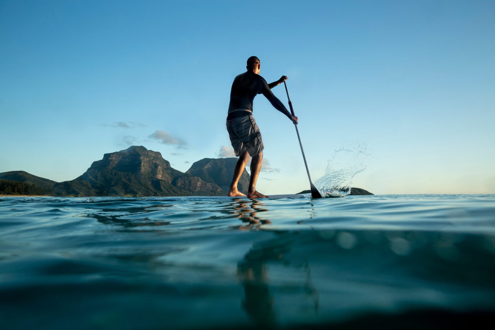 Stand Up paddle boarding on Calm ocean water, young adult surfer male standing on a surfboard and using a paddle, water surface rear view, Mount Gower and Lidgbird view, Lord Howe Island,travel, new south wales, tasman sea, low angle, water view,